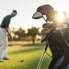 A photo of senior man and woman at golf course. Focus is on golf bag. They are playing on sunny day.